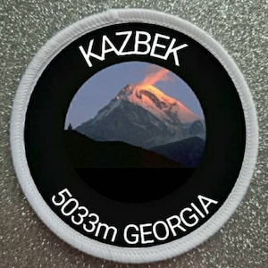 May include: White round patch with black border and a photo of a snow-capped mountain peak. The text "KAZBEK 5033m GEORGIA" is printed in white on the patch.