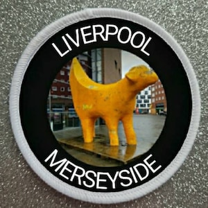 Puede incluir: Parche redondo con las palabras "LIVERPOOL MERSEYSIDE" en blanco sobre un fondo negro. El parche presenta una escultura amarilla de un perro en el centro, sobre un fondo exterior borroso. Borde blanco.
