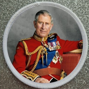 May include: A round embroidered patch featuring a portrait of a man in a red military uniform with gold trim and medals. He is wearing a blue sash and a gold belt. The background is a grey wall.