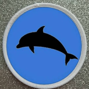 May include: A white circular patch with a blue background and a black silhouette of a dolphin jumping out of the water.