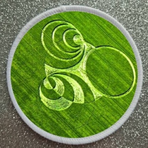 Könnte beinhalten: Ein weißer Patch mit einem grünen Kreis in der Mitte. Der Kreis zeigt ein komplexes Muster aus konzentrischen Kreisen und Linien, das ein geometrisches Design erzeugt.