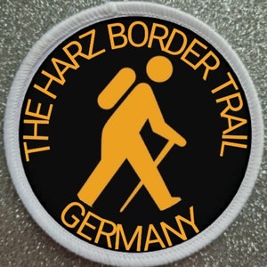 May include: A black and white circular patch with a white border. The patch features a yellow hiker with a backpack and walking stick. The text "THE HARZ BORDER TRAIL GERMANY" is written in yellow around the hiker.