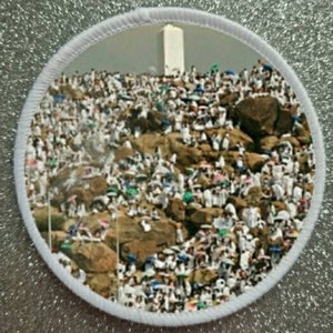 Puede incluir: Un parche blanco con una imagen circular de una gran multitud de personas vestidas de blanco reunidas en una ladera rocosa. Una estructura blanca alta es visible en el fondo.