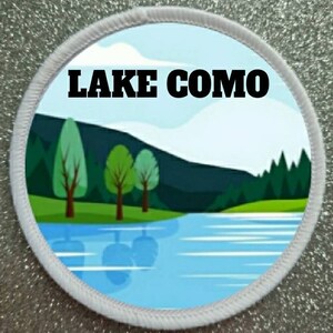 Könnte beinhalten: Ein runder Aufnäher mit einer Landschaftsillustration des Comer Sees. Das Design zeigt einen blauen See, grüne Bäume und eine Bergkette unter einem hellblauen Himmel. Der Text "LAKE COMO" ist in fetten schwarzen Buchstaben.