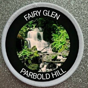 May include: White round patch with black border and text "Fairy Glen Parbold Hill". The center of the patch features a photo of a waterfall in a lush green forest.