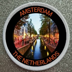 May include: A white circular patch with black and orange text. The text reads "Amsterdam The Netherlands". The centre of the patch features a photo of a canal in Amsterdam.