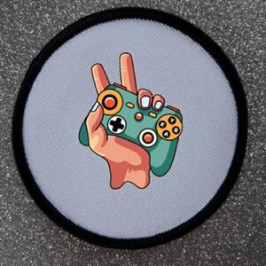 May include: A white round patch with a black border features a cartoon hand holding a green and yellow video game controller. The hand is making a peace sign with two fingers.