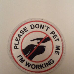 Peut inclure: Un patch rond blanc avec des coutures rouges. Le patch a un cercle rouge avec une ligne rouge diagonale qui le traverse. Le texte "PLEASE DON'T PET ME I'M WORKING" est écrit autour du cercle. À l'intérieur du cercle se trouve une silhouette noire de la tête d'un chien avec une main qui tend la main pour le caresser.