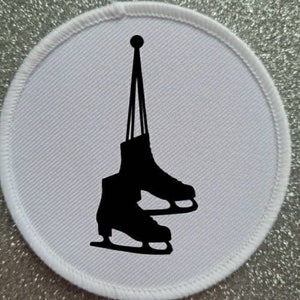 Puede incluir: Un parche redondo blanco con una silueta negra de un par de patines de hielo colgando de un gancho.
