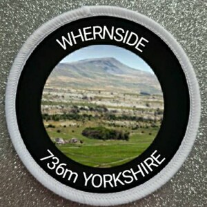Może przedstawiać: Okrągła naszywka w kolorze białym i czarnym z tekstem "Whernside 736m Yorkshire" i zdjęciem zielonego pola z górą w tle.