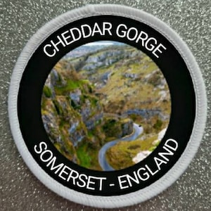 May include: White round patch with black text and a black circle in the centre. The text reads "Cheddar Gorge Somerset - England". The circle contains a photo of a winding road through a green valley.