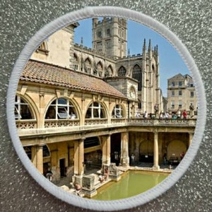 May include: A circular image of the Roman Baths in Bath, England. The image shows the exterior of the baths, with the Bath Abbey in the background. The baths are made of stone and have a green pool of water in the centre.