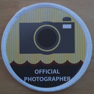 Könnte beinhalten: Ein runder weißer Aufnäher mit einem gelben und braunen gestreiften Hintergrund. Ein schwarzes Kamerasymbol befindet sich mittig auf dem Aufnäher. Der Text "OFFICIAL PHOTOGRAPHER" ist in weiß auf schwarzem Hintergrund am unteren Rand des Aufnähers aufgedruckt.