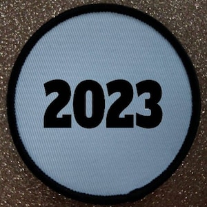 Op de afbeelding: Een ronde witte patch met een zwarte rand en het jaar 2023 in het zwart gedrukt.