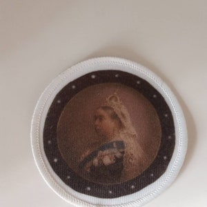 May include: Circular patch with a portrait of a historical figure in profile, wearing a crown and formal attire. The portrait is framed by a dark brown circle with white dots, and a white stitched edge.