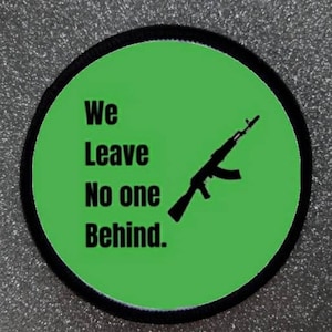 Può includere: Toppa rotonda con centro verde e bordo nero. Il centro verde presenta il testo "We Leave No one Behind" in nero, insieme a una silhouette nera di un fucile.
