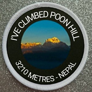 Może przedstawiać: Okrągła biała naszywka z czarnym tekstem, który brzmi "I've Climbed Poon Hill 3210 Metres - Nepal". Naszywka zawiera zdjęcie pasma górskiego z niebieskim niebem i chmurami w tle.