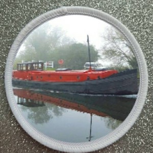 May include: A red and black boat is moored in a canal with a reflection of the boat in the water. The boat is in a misty setting with trees in the background.