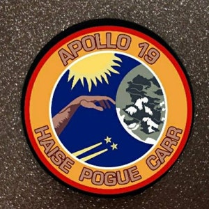 Pode incluir: Um remendo redondo, laranja, vermelho e preto com o texto "Apollo 19" no topo. O remendo apresenta uma imagem estilizada de uma mão que alcança uma lua crescente com um sol ao fundo. O texto "Haise Pogue Carr" está escrito ao redor da parte inferior do remendo.