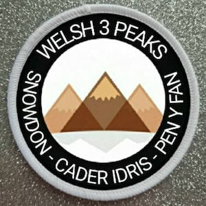 May include: White round patch with black text and a graphic of three mountains. The text reads "Welsh 3 Peaks Snowdon - Cader Idris - Pen y Fan".