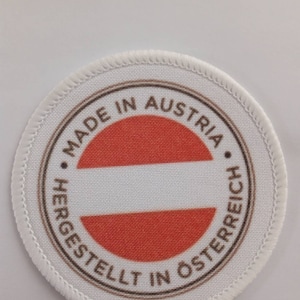 May include: A white round patch with brown text that reads "Made in Austria - Hergestellt in Österreich" and a red and white Austrian flag in the center.