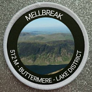 May include: Circular patch with a landscape image of a mountain and lake. The patch has a black border with white text that reads "MELLBREAK", "512 M - BUTTERMERE - LAKE DISTRICT". The patch has a white stitched border and is set against a glittery background.