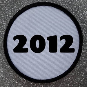 Könnte beinhalten: Runder weißer Patch mit schwarzem Rand und der Jahreszahl 2012 in Schwarz gedruckt.