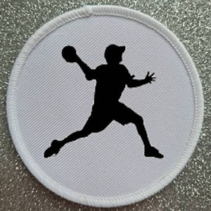 May include: A circular patch featuring a white background with a black silhouette of a person throwing a ball. The patch has a white stitched border, possibly an emblem or logo.