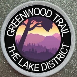 May include: A white circular patch with black text that reads "Greenwood Trail The Lake District" around a black circle with a purple and orange sunset scene with a silhouette of a tree and mountains.