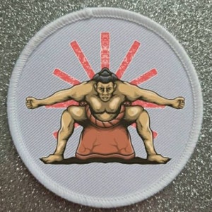 May include: A white round patch with a red sunburst design and a cartoon image of a sumo wrestler in a crouching position. The wrestler is wearing a traditional loincloth and has a serious expression on his face.