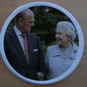 May include: A round, white-bordered patch with a photo of Queen Elizabeth II and Prince Philip. The Queen is wearing a light blue dress and a pearl necklace. Prince Philip is wearing a dark suit and a red tie.