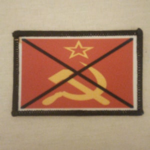 Pode incluir: Um remendo vermelho com um X preto sobre um martelo e uma foice amarelos e uma estrela amarela. O remendo tem uma borda preta.