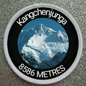 May include: White round patch with black border and white text. The text reads "Kangchenjunga 8586 METRES". The patch features a photo of a snow-capped mountain.