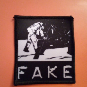 Könnte beinhalten: Schwarz-weiß gestickter Patch mit einem Astronauten auf dem Mond und dem Wort "FAKE" darunter.