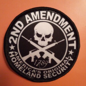 Könnte beinhalten: Ein runder schwarzer und weißer Patch mit dem Text "2nd Amendment" in einem Kreis um einen Totenkopf und gekreuzte Knochen. Der Text "America's Original Homeland Security" befindet sich unter dem Totenkopf und den gekreuzten Knochen. Das Jahr "1789" ist in Kursivschrift unter den gekreuzten Knochen geschrieben.