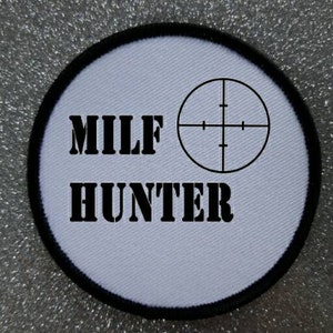Op de afbeelding: Ronde witte patch met zwarte tekst en een doelwitsymbool. De tekst luidt "MILF HUNTER".