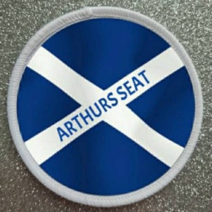 May include: White round patch with a blue and white Scottish flag design. The text "ARTHUR'S SEAT" is written in black across the white diagonal stripe.