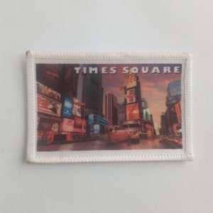 May include: A white fabric patch with a colourful image of Times Square in New York City. The patch has a white border with a stitched edge.