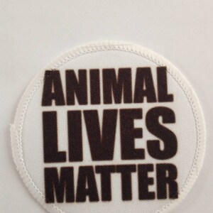 May include: A circular white patch with the words "ANIMAL LIVES MATTER" in dark brown block letters. The patch has a white stitched border. The background is plain white.