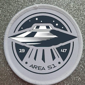 Puede incluir: Un parche redondo blanco con un diseño de OVNI en blanco y negro. El OVNI está volando sobre un círculo negro con el texto "AREA 51" y los números "19" y "47" a cada lado.