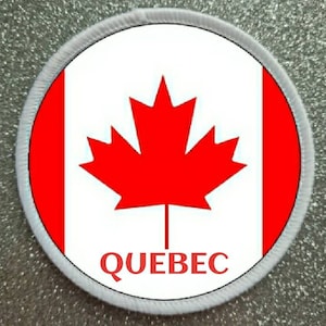 Può includere: Una toppa circolare bianca e rossa con una foglia d'acero rossa e la parola "QUEBEC" in testo nero.