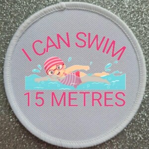 May include: White round patch with pink text that reads "I can swim 15 metres". A cartoon illustration of a girl wearing a pink swim cap and goggles swimming in blue water.