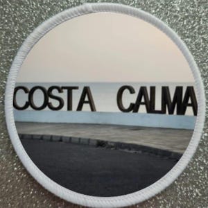 May include: A white round patch with a black border and the words "COSTA CALMA" in black letters against a light blue and white background.