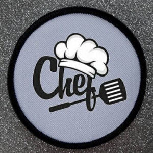 Pode incluir: Um chapéu de cozinheiro branco com uma espátula preta e a palavra "Chef" em uma fonte estilizada sobre um fundo cinza. O design está em um patch redondo com borda preta.
