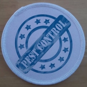 Op de afbeelding: Ronde witte stoffen patch met een blauwe cirkel en de tekst "Pest Control" in een diagonale blauwe stempel. De cirkel is omgeven door acht blauwe sterren.