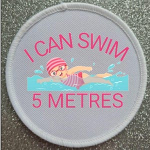 Puede incluir: Un parche redondo blanco con texto rosa que dice "I CAN SWIM 5 METRES". Una ilustración de dibujos animados de una persona nadando en agua azul está en el centro del parche.