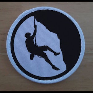 Puede incluir: Un parche redondo blanco y negro con la silueta de una persona escalando. La persona está escalando una pared de roca negra.