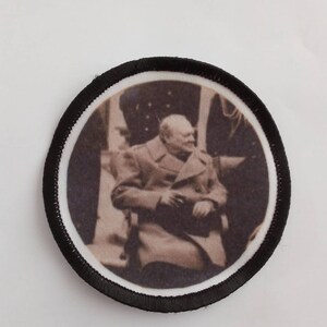 May include: A round black and white patch with a black border. The patch features a black and white photo of a man in a suit and tie. The man is sitting down and looking to the side.