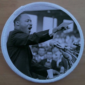 Può includere: Foto in bianco e nero di Martin Luther King Jr. che tiene un discorso. Indossa un abito e una cravatta e indica a destra. Ci sono microfoni davanti a lui e una folla di persone dietro di lui.