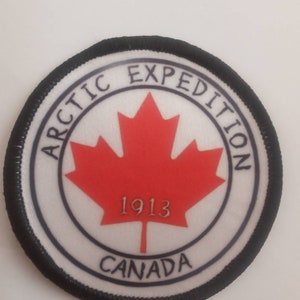 Op de afbeelding: Een witte ronde patch met een zwarte rand. De patch heeft een rood esdoornblad met de tekst "ARCTIC EXPEDITION 1913 CANADA" in zwart.